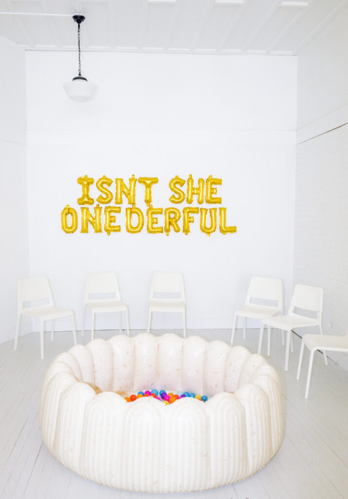 Gold balloons on wall that spell "isnt she onederful" and a white blow up ball pit for a garden party themed birthday. 