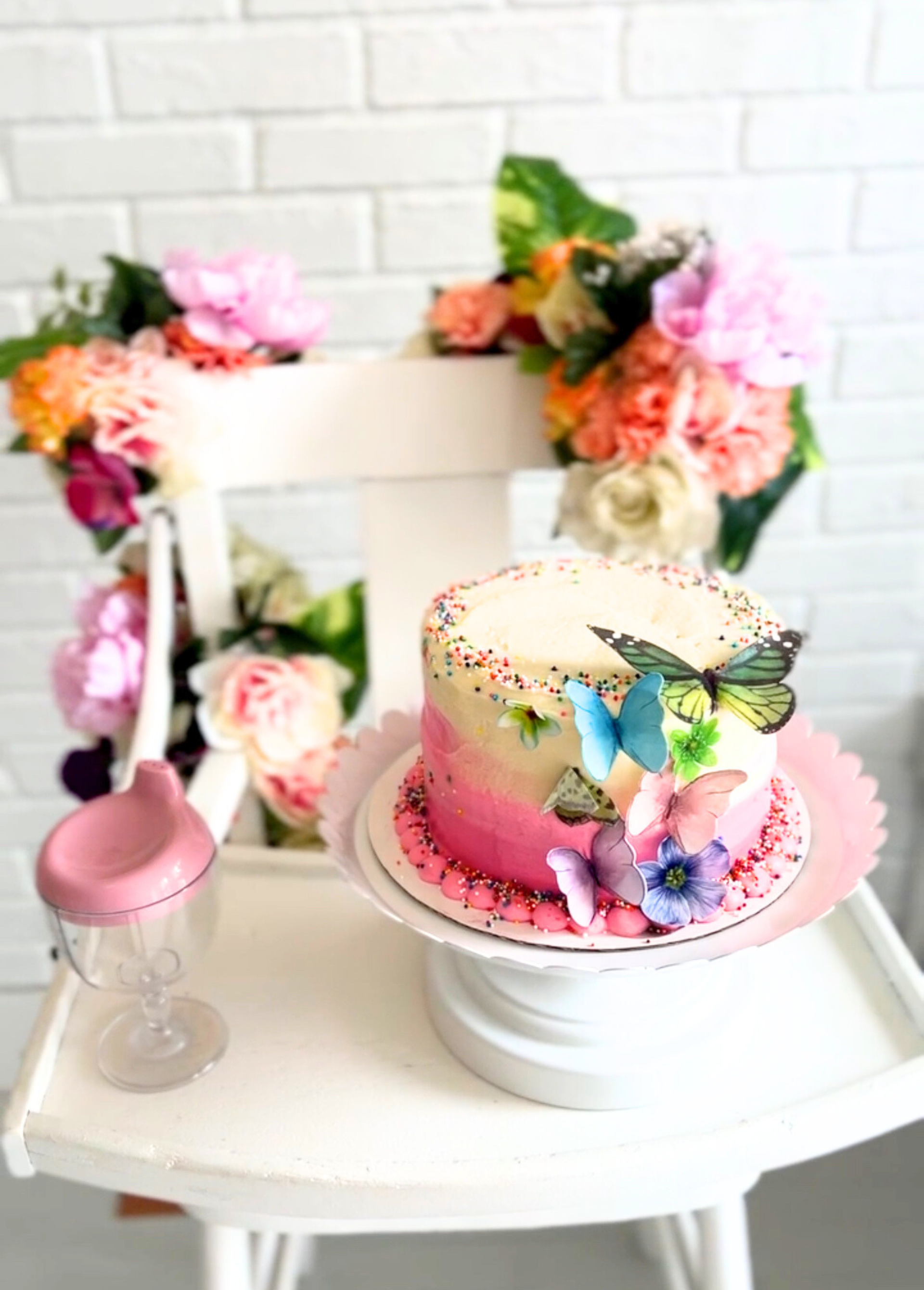 Colorful smash cake with edible butterflies sitting on a beautifully decorated white highchair with flowers adorning it.