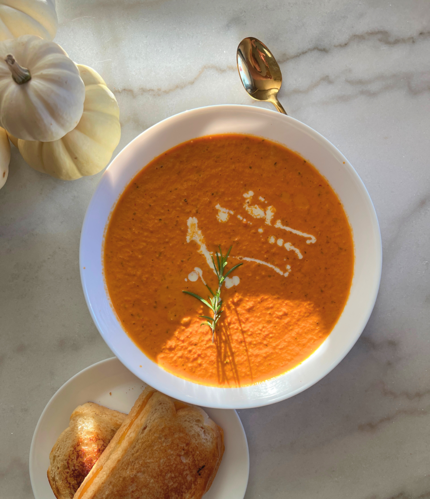 creamy roasted tomato soup in a white bowl with a rosemary sprig on top sitting next to grilled cheese sandwich and some what pumpkins on a white marble top dining room table.