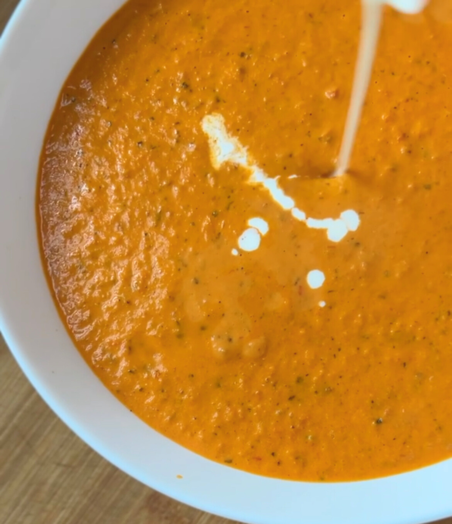 creamy roasted tomato soup in a white bowl with heavy cream being drizzled on top.