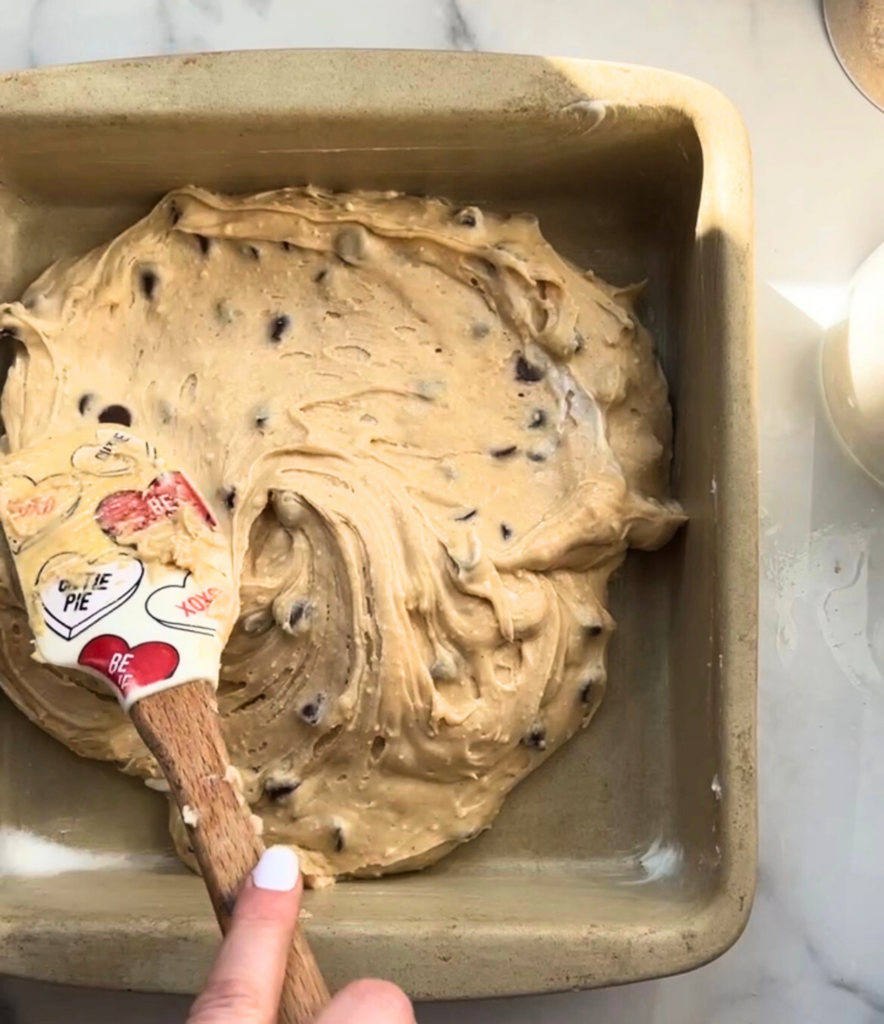 Spreading chocolate chip cookie cake batter into a prepared baking pan