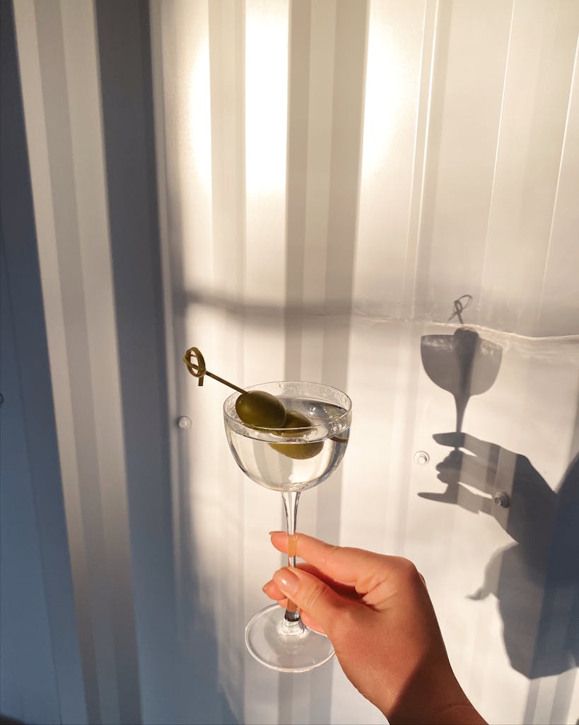 Close-up of a hand holding a martini glass with olives against a light backdrop.