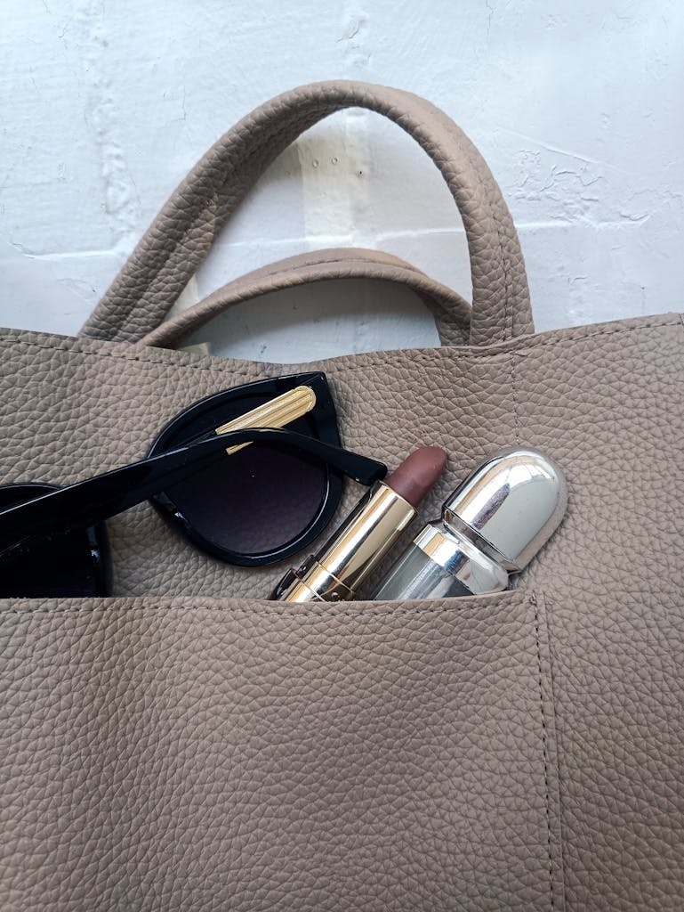 Close-up view of a handbag containing sunglasses, lipstick, and perfume on a textured surface.